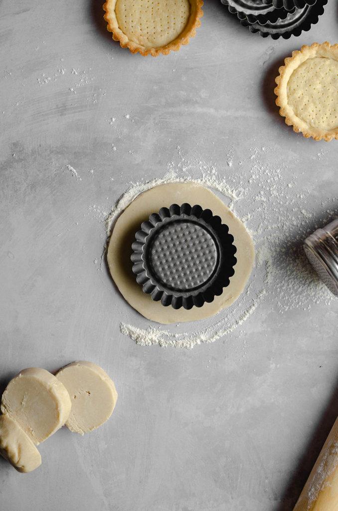 Step one, how to form a shell for vegan custard tarts. Rolled out crust sitting underneath a mini tart tin. Dusted flour all over the surface.