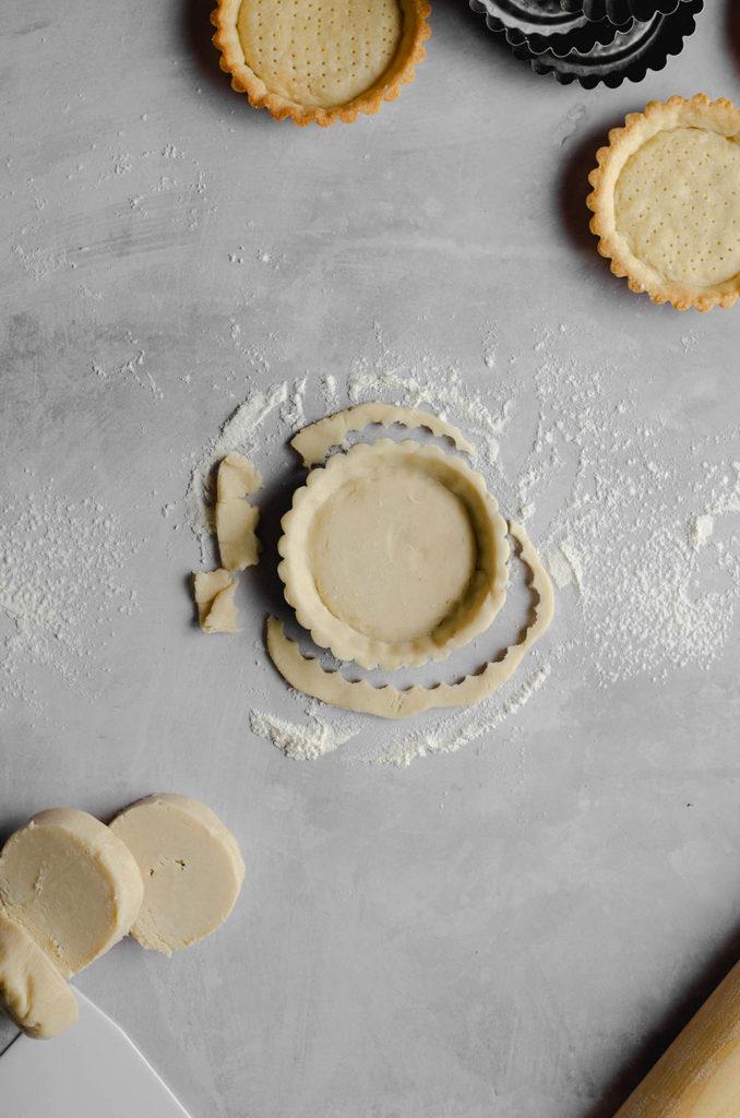 Step four, how to form a shell for vegan custard tarts. Tart shell dough in the the tart tin, with scraps of dough around it. Flour dusted all over the surface.