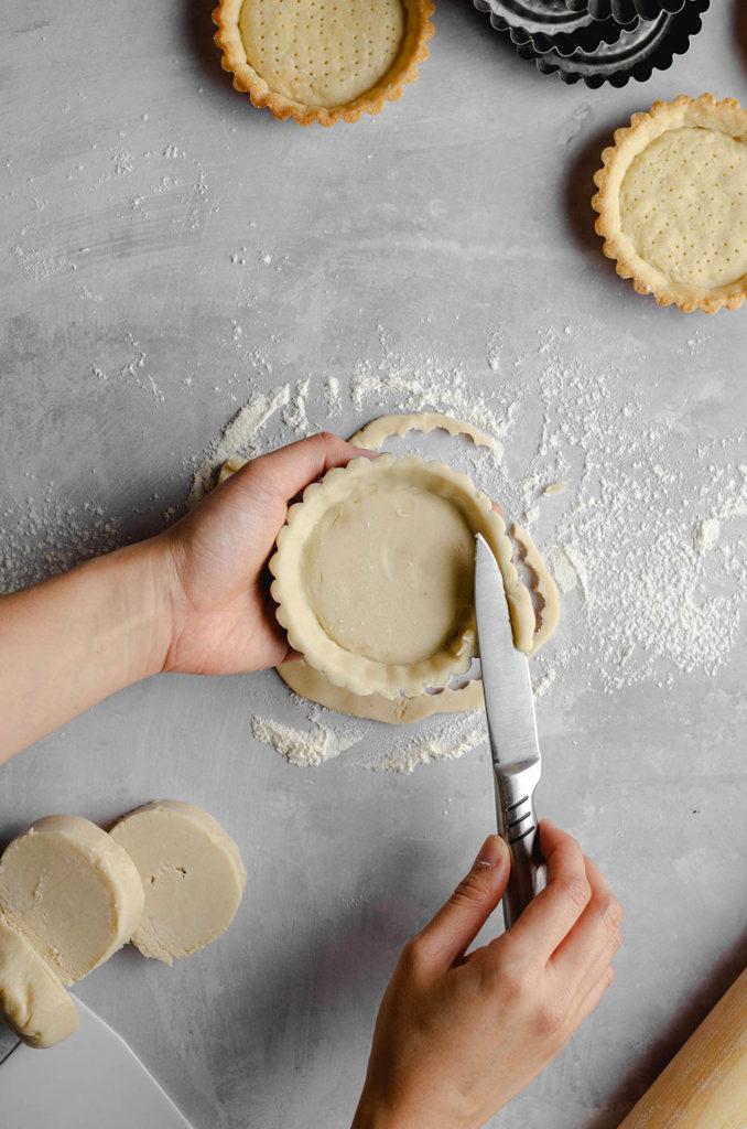 Step five, how to form a shell for vegan custard tarts. Left hand holds the tart tin, and right hand is holding a pairing knife. Cutting remaining dough and to level out the tart shell.
