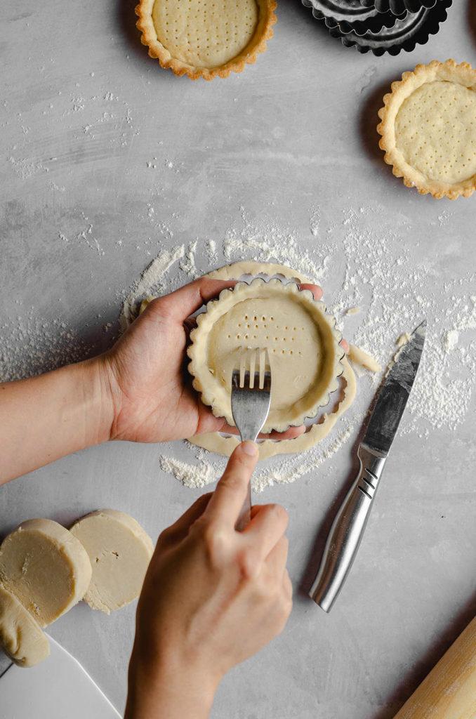 Step six, how to form a shell for vegan custard tarts. Left hand holds the tart tin, and right hand is holding a fork. Lightly pricking holes on the bottom of the shell.