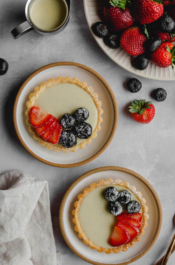 Top view, two vegan custard tarts on small plates, topped with blueberries, strawberries and powdered sugar. Plate full of berries sitting on the top right.