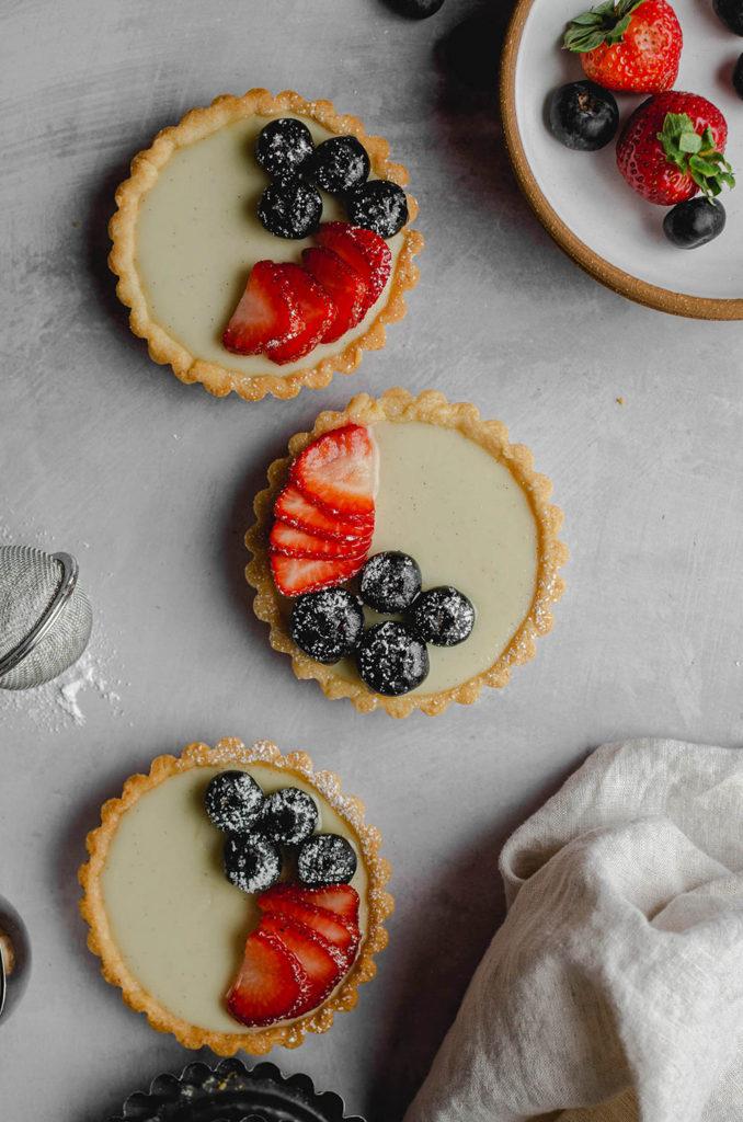 Top view, three tarts topped with blueberries, strawberries, and powdered sugar, sitting over a surface. Plate of berries on the top right.