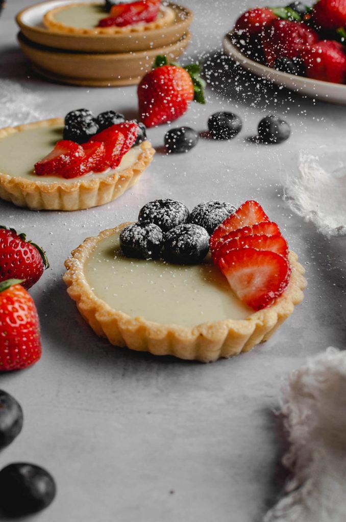 45 degree angle, a single custard in the middle, topped with blueberries and strawberries. Motion of powdered sugar being dusted over top. Extra custard tarts and berries in the back and out of focus.