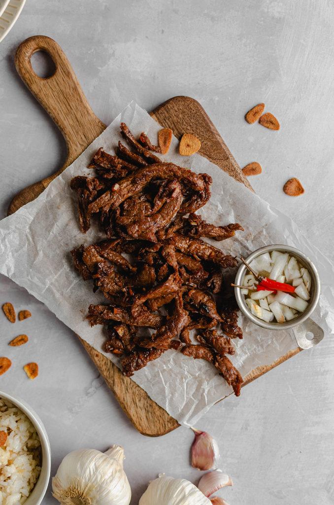 Top view, vegan tapa meat laid on a wooden chopping board and over a crumpled parchment paper. A small bowl of spicy vinegar on the right side, slightly out of frame. Bowl of rice on bottom left with bulbs and pieces of garlic, out of frame. Garlic chips sprinkled over the scene.
