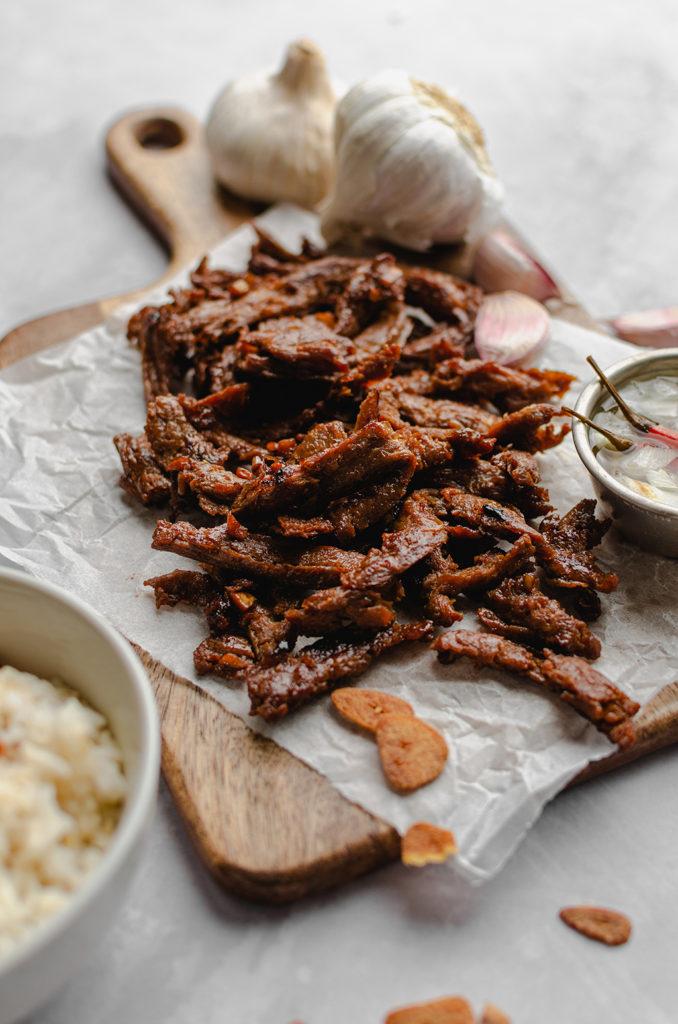 Vegan tapa meat laid on a wooden chopping board and over a crumpled parchment paper. A small bowl of spicy vinegar on the right side, slightly out of frame. Bowl of rice in the front and on the bottom left, out of focused. Bulbs and pieces of garlic in the back and out of focus.