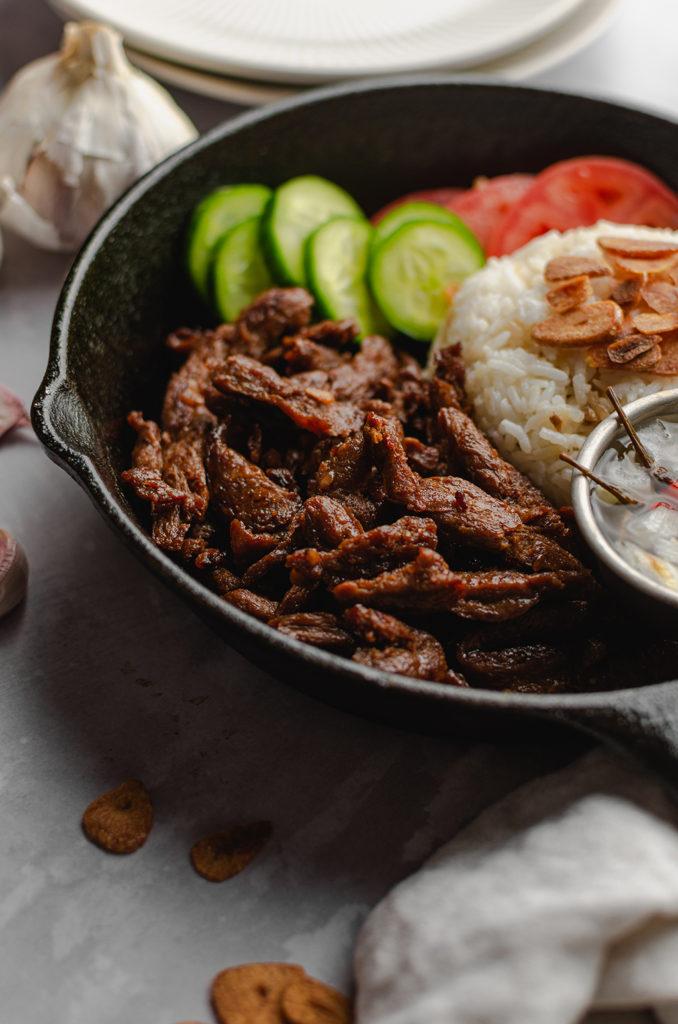 45 degree angle, cast-iron pan filled with vegan meat, garlic rice, cucumbers, tomatoes, and a bowl of spicy vinegar. Vegan meat is in focus.