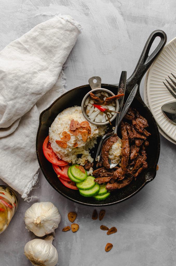 Top view of a cast-iron pan filled with garlic rice, vegan meat, cucumbers, tomatoes, and bowl of spicy vinegar. Fork and spoon in the cast-iron pan set as if the food was being eaten.
Bottle of spicy vinegar out of frame on bottom left, along with two garlic bulbs. Fried garlic chips sprinkled on the bottom of the cast-iron. Plates and form and spoon out of frame on the right side.