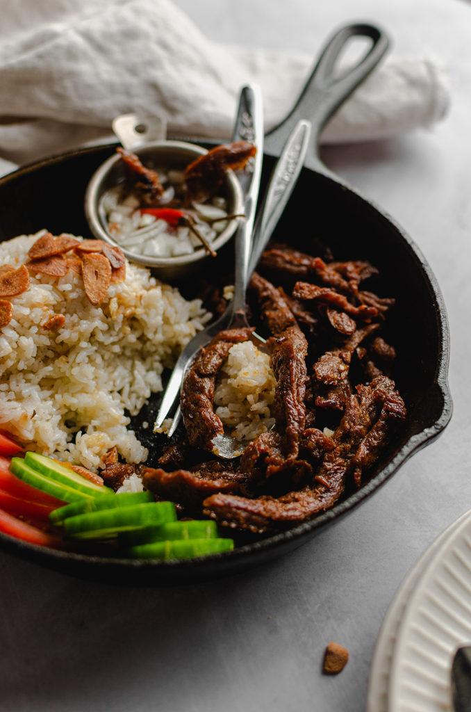 45 degree angle a cast-iron pan filled with garlic rice, vegan meat, cucumbers, tomatoes, and bowl of spicy vinegar. Fork and spoon in the cast-iron pan set as if the food was being eaten.