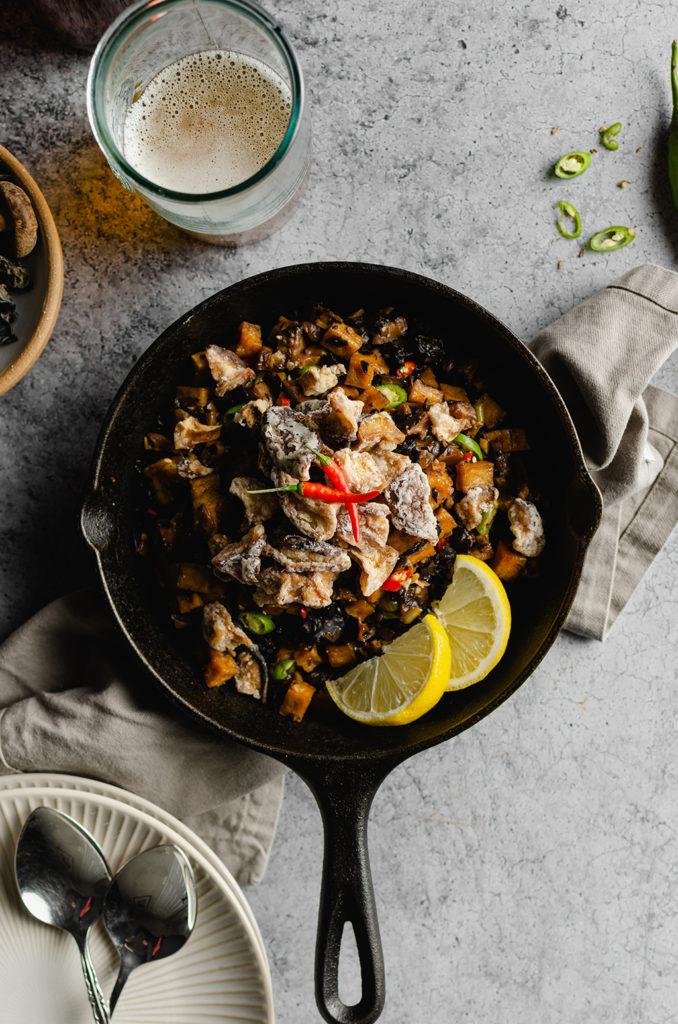 Top view of a cast-iron sitting over a gray napkin. Cast-iron is filled with vegan Filipino sisig, topped with two red Thai chili peppers and two slices of lemon on the side.