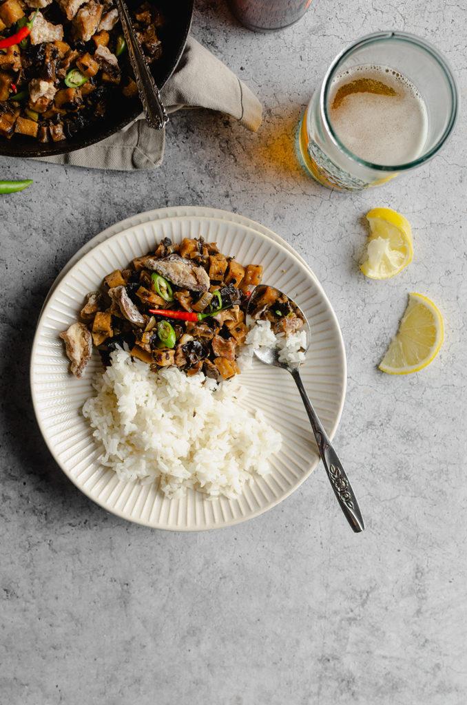 Top view of a plate half filled with the vegan Filipino sisig and rice. A spoon on the plate used as if someone was eating. Lemon slices on the center right. Glass of beer on the top right. More sisig at the top left.