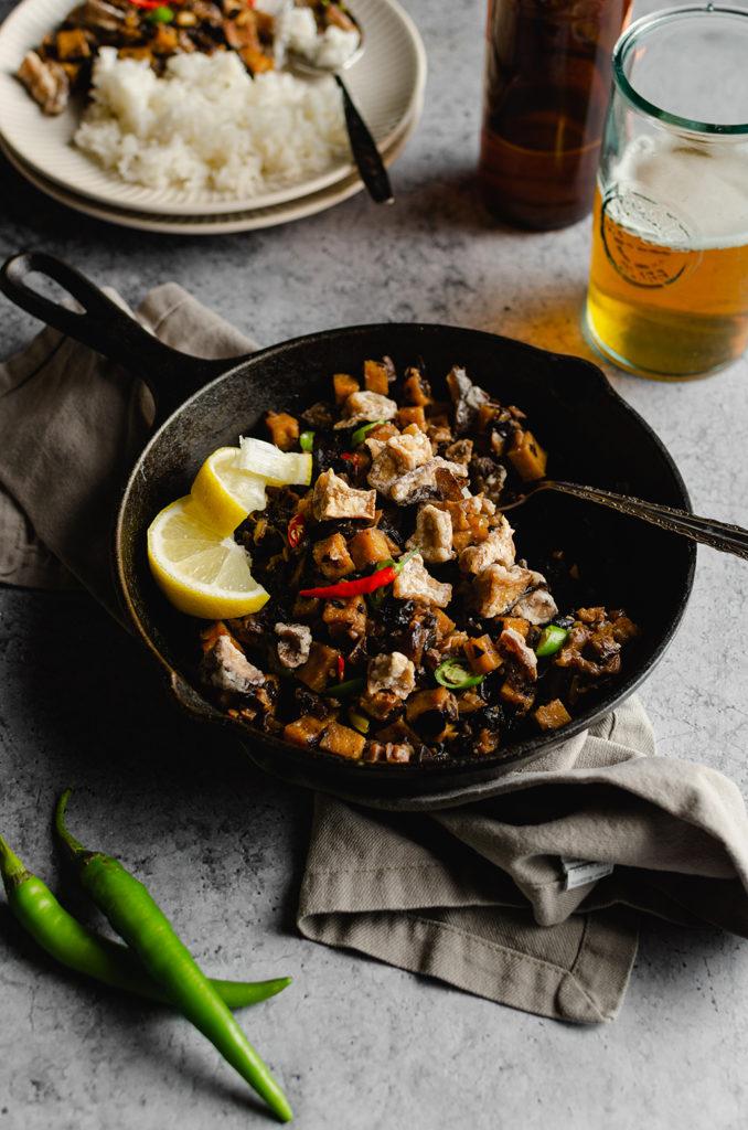 45 degree angle of a cast-iron sitting over a gray napkin. Cast-iron is filled with vegan Filipino sisig, topped with two red Thai chili peppers and two slices of lemon on the left side. A spoon in the cast-iron to show someone was eating from it. Bottom left are green finger chilis. Top right is glass of beer. In the back, a plate of sisig and rice, and out of focus.