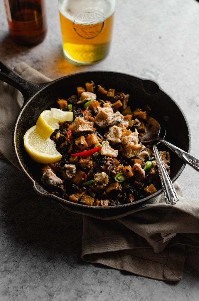 45 degree angle of a cast-iron sitting over a gray napkin. Cast-iron is filled with vegan Filipino sisig, and two slices of lemon on the left side. Two spoons resting in the cast-iron to show someone was eating from it. In the back, a bottle and glass of beer out of focus.