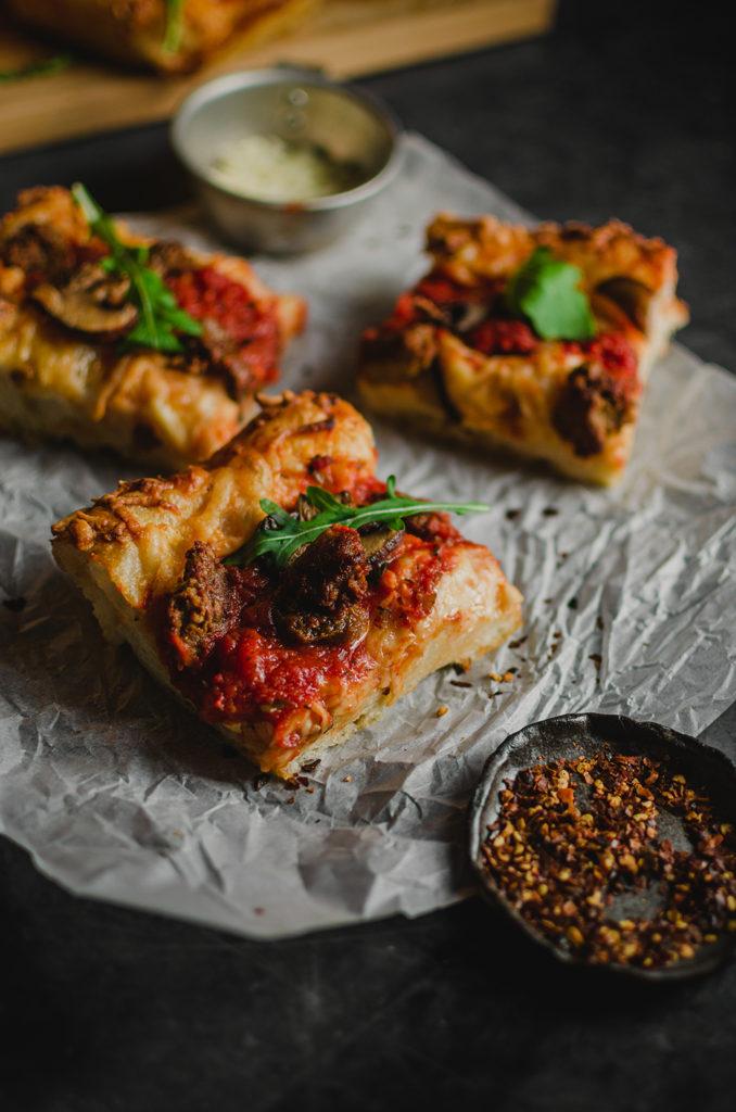45 degree angle, a piece of pizza in the center and in focus. Set on top of a piece of parchment paper. A small plate of chili flakes in the foreground and out of focus. Two pieces of pizza behind and out of focus.