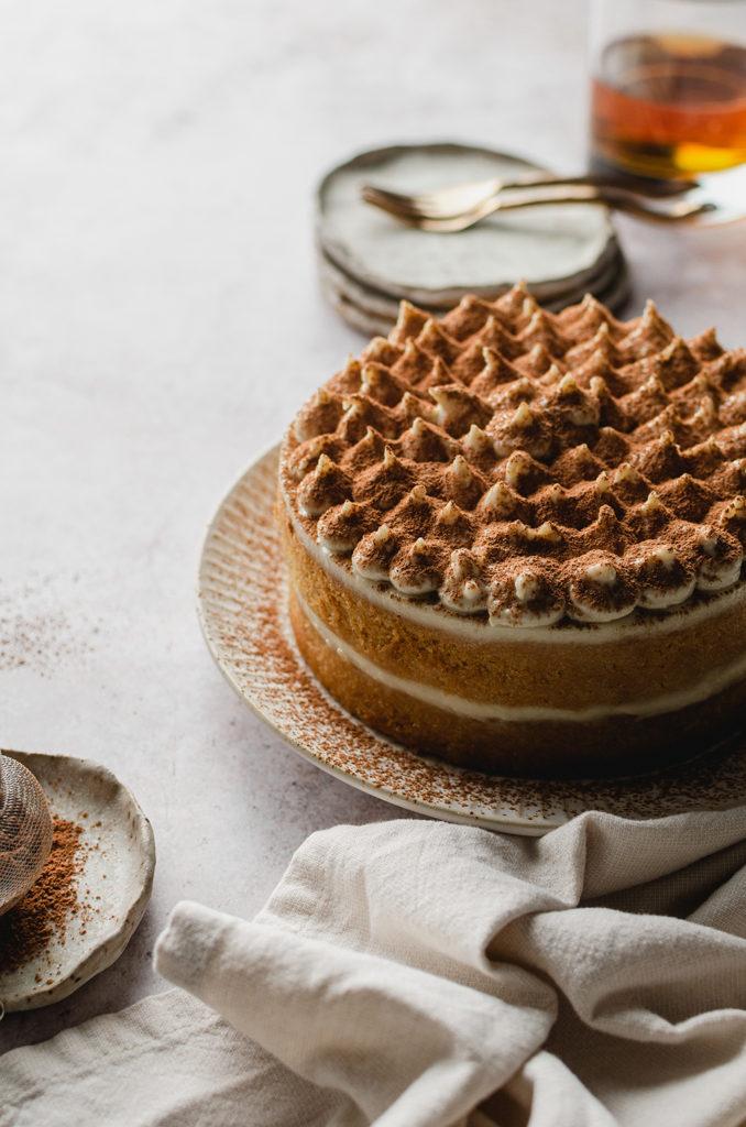 Whole tiramisu cake with dollops of cream on top. Sifted cocoa powder off to the left side to show it was freshly dusted with cocoa powder. Plates, forks, and a glass of iced tea out of focus in the background.