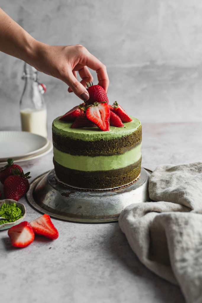 Front view of the whole matcha cake with hands topping it off with strawberries