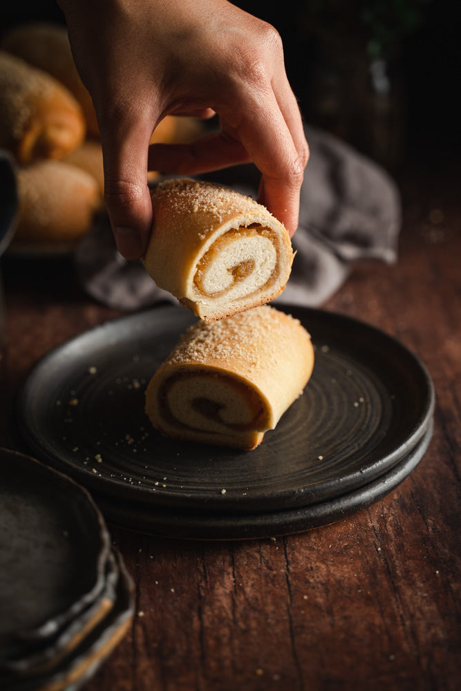 A front view of a vegan Spanish bread cut in half, revealing the swirly center with the filling. A hand taking one half.