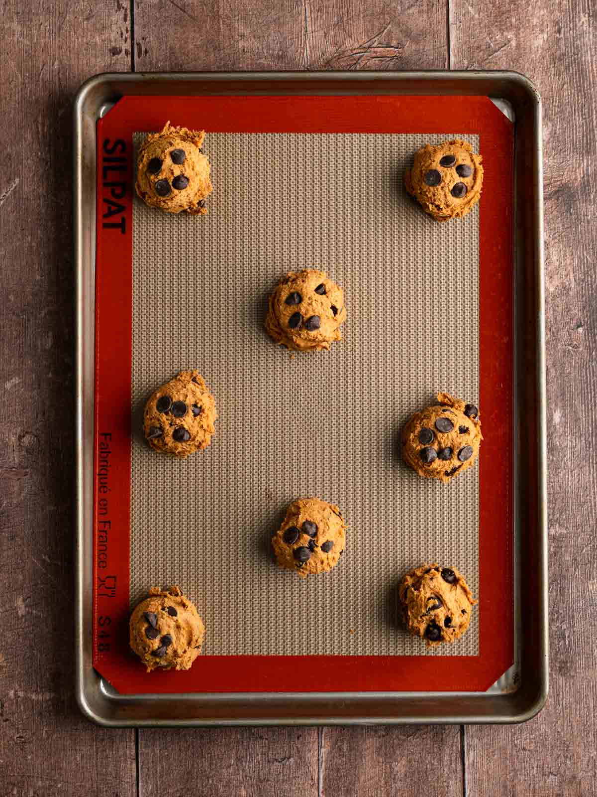 Eight scoops of raw cookie dough on a lined baking sheet.