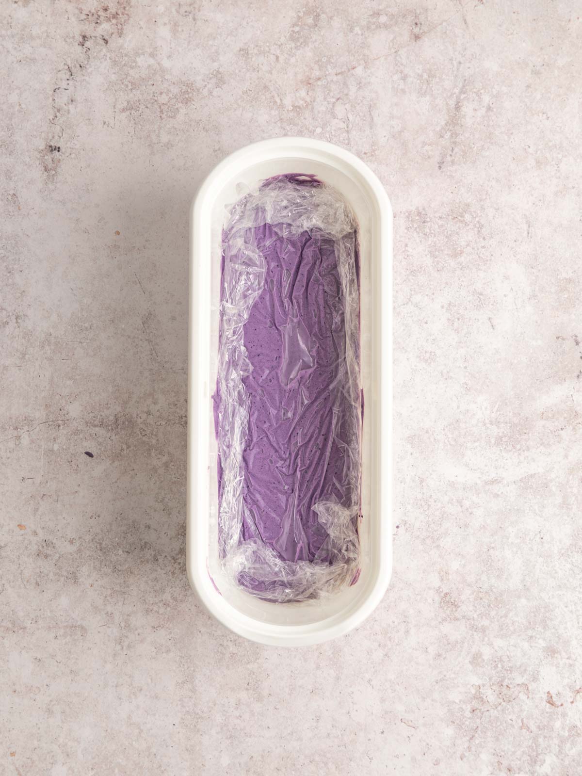Vegan ube ice cream poured in a freezer-safe container, and cling film set over the surface.