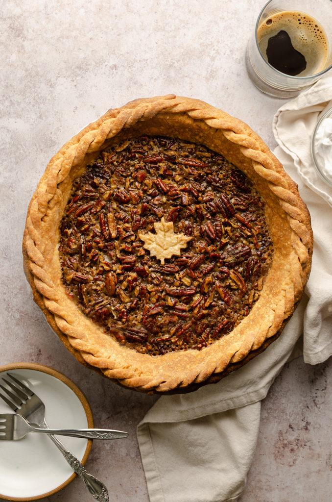 Top view of the vegan maple bourbon pecan pie, with a pie crust maple leaf decoration in the middle. Bottom left a plate with forks. Top right a glass of black coffee and a bowl of whipped cream.