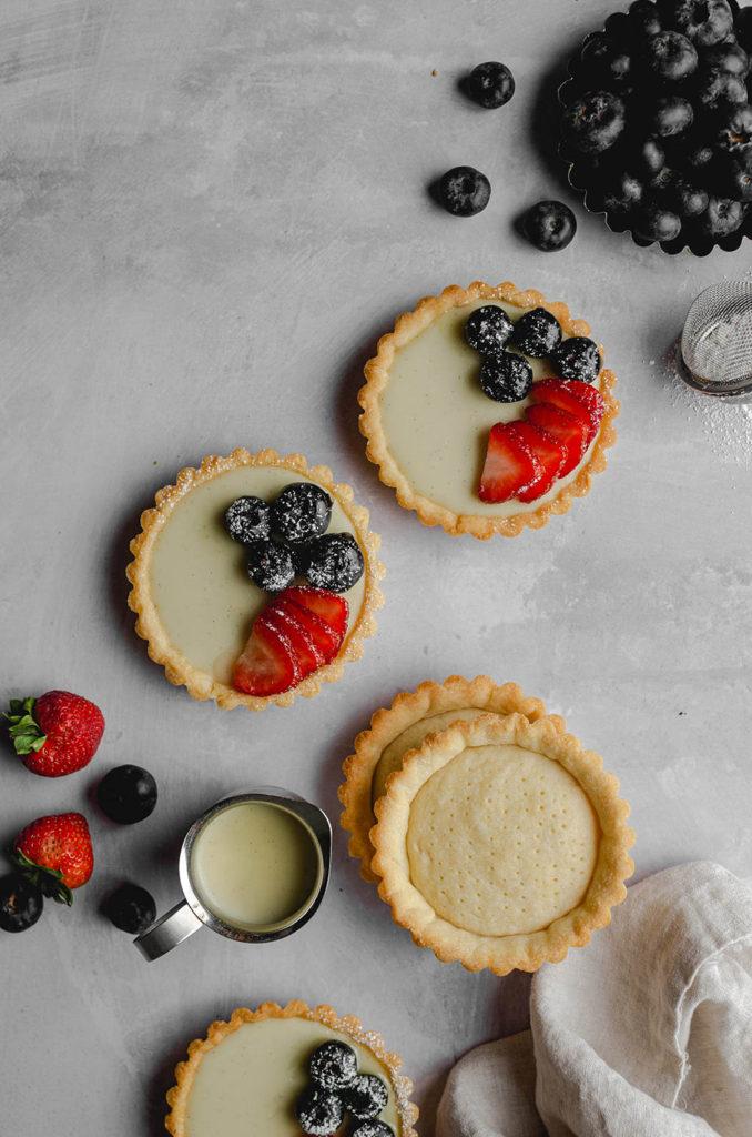 Top view, two vegan custard tarts in the middle, topped with blueberries, strawberries and powdered sugar. Next to it are empty tart shells and a mini pitcher full of custard. Extra berries surrounding the custards.