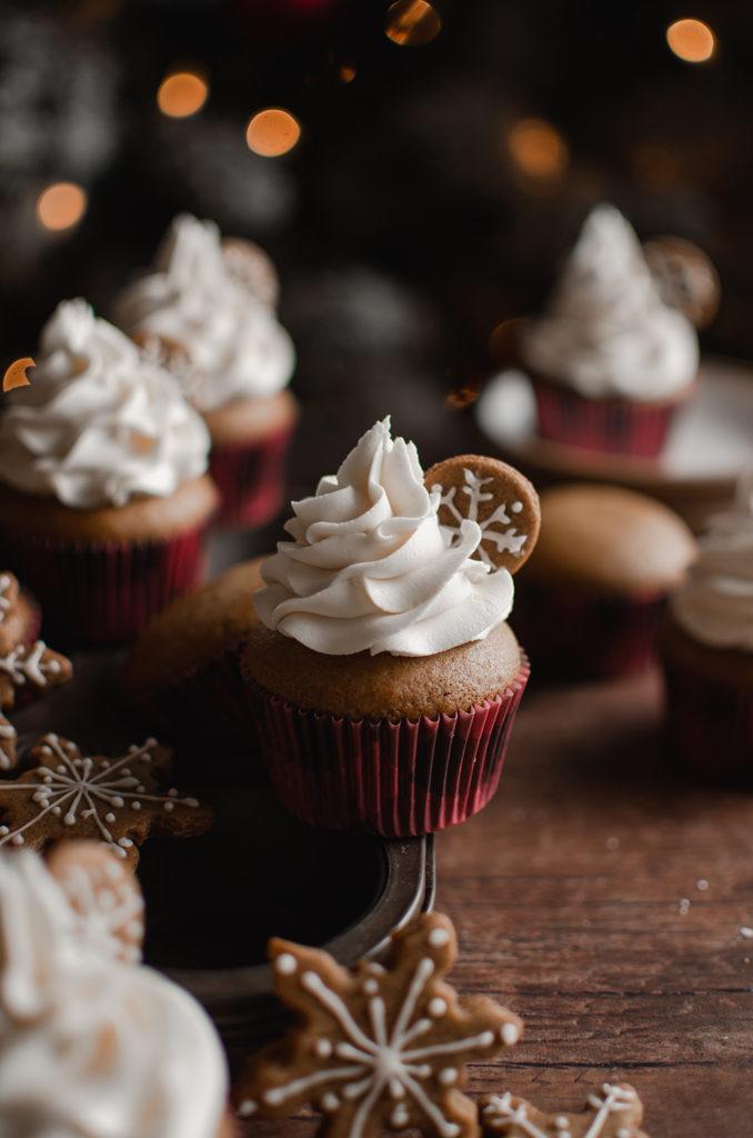 45 degree angle shot, a single vegan gingerbread cupcake in center and in focus. Topped with swirl of buttercream and a small cookie inserted. Gingerbread cupcakes and cookies surrounding  it and out of focus.