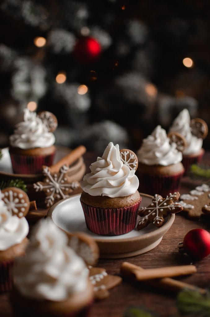 45 degree angle shot, a single vegan gingerbread cupcake resting on a plate with a snowflake cookie next to it. Topped with swirl of buttercream and a small cookie inserted. Gingerbread cupcakes and cookies in the background and foreground and out of focus.