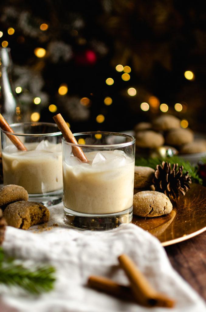 Front shot of two glasses of creamy vegan eggnog with one cinnamon sticks in each glass. Placed on top of a gray napkin and a gold plate charger. Cookies spread around the glasses. Christmas lights blurred in the background.