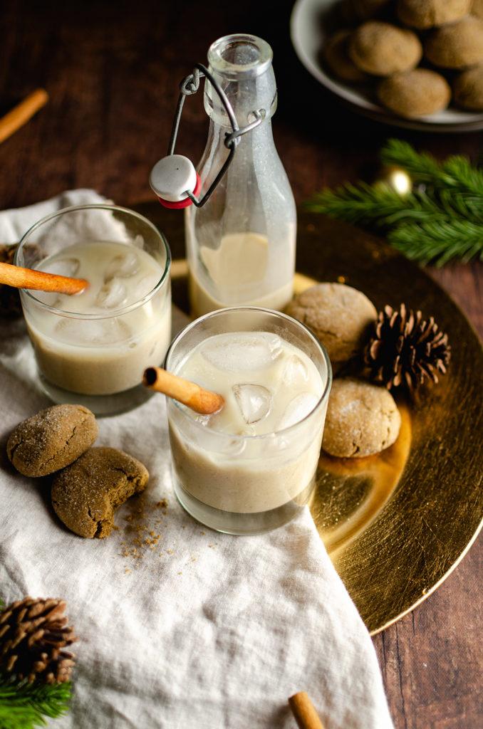 45 degree angle of a two glasses and a bottle filled with creamy vegan eggnog, with one cinnamon stick in each glass. Placed on top of a gray napkin and a gold plate charger. Cookies surrounding the glasses. Plate of cookies and green pine tree branches in the back, top right, out of focus.