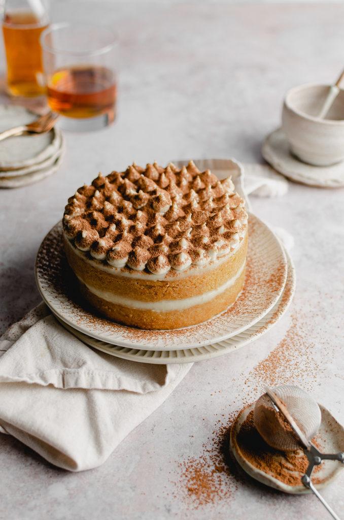 Whole layer cake in the center, dolloped with fresh cream. Sifted cocoa powder on the bottom right. Bottle of tea, plates, and forks out of focus in the back.