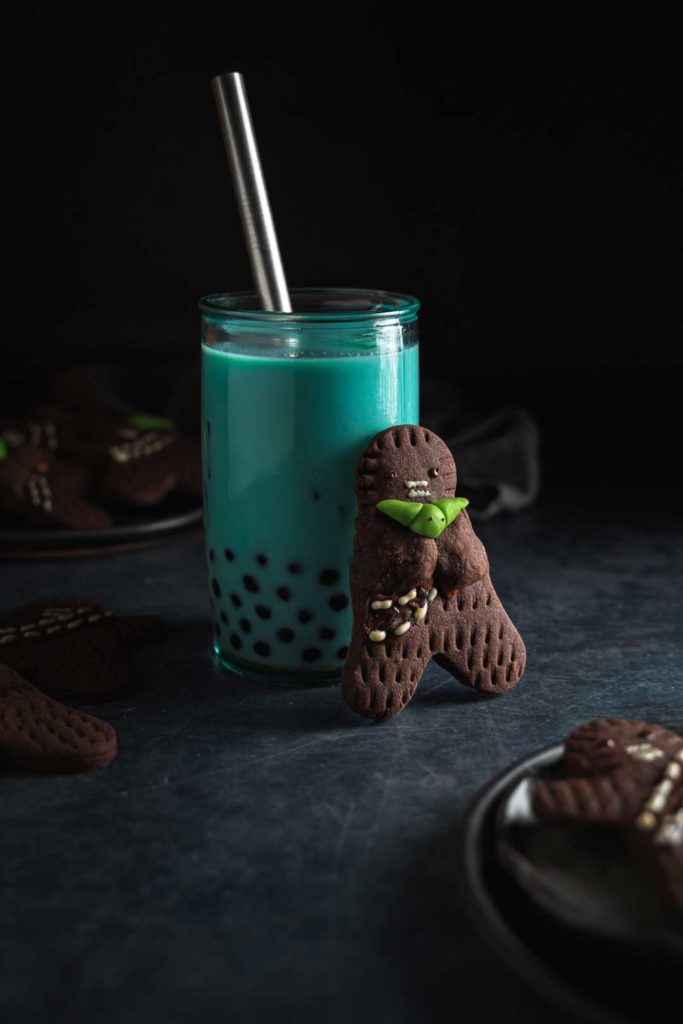 A glass of butterfly pea milk tea and a  wookie cookie standing next to the cup.