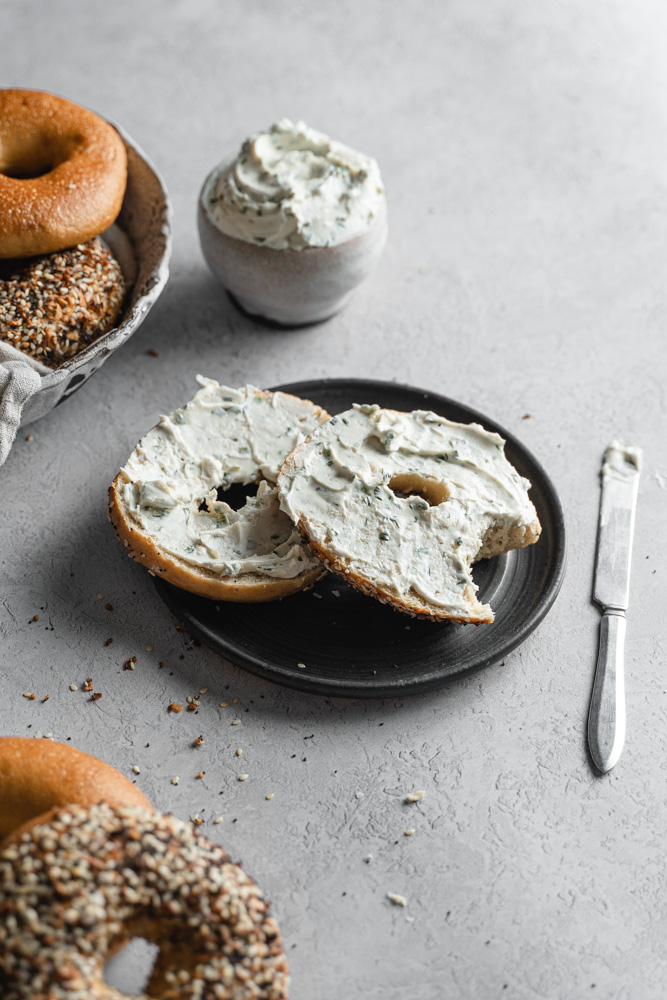 A New York-Style Bagel on a plate, cut in half, some cream cheese spread and a bit taken out of it.